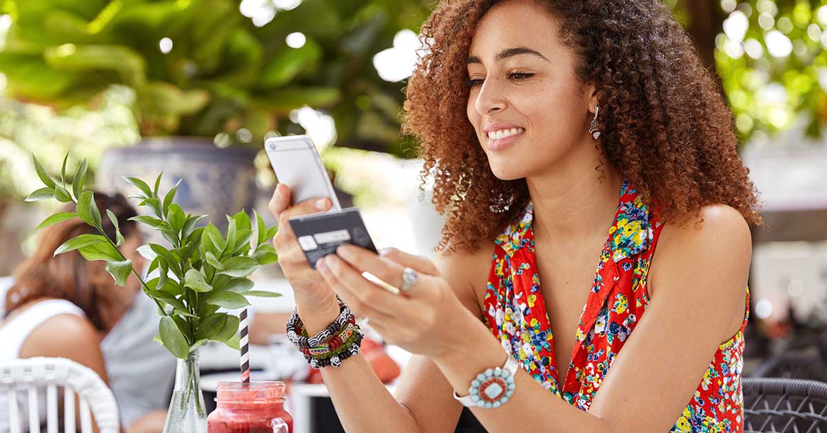 photo of a woman in her 20s looking at her credit card while holding a mobile phone in her other hand