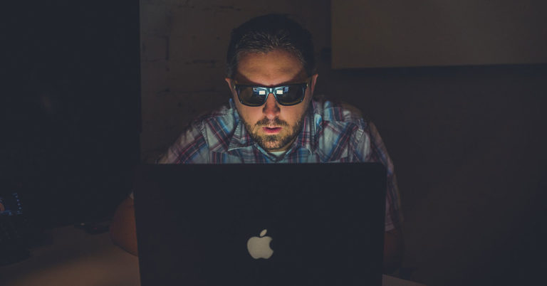 there are practical ways to protect your online identity and profiles; photo of man wearing sunglasses in dark room typing on computer
