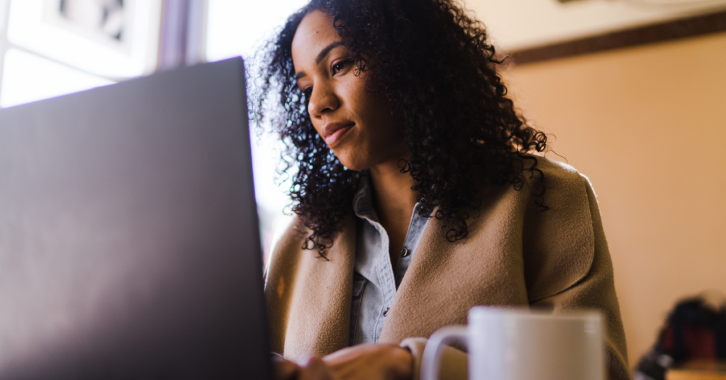 Woman with curly hair in a tan coat uses a laptop at a cafe, with a mug in the foreground. | Honor Credit Union