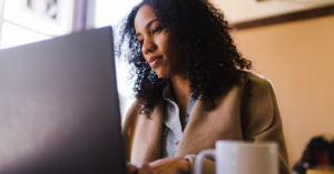 Woman with curly hair in a tan coat uses a laptop at a cafe, with a mug in the foreground. | Honor Credit Union