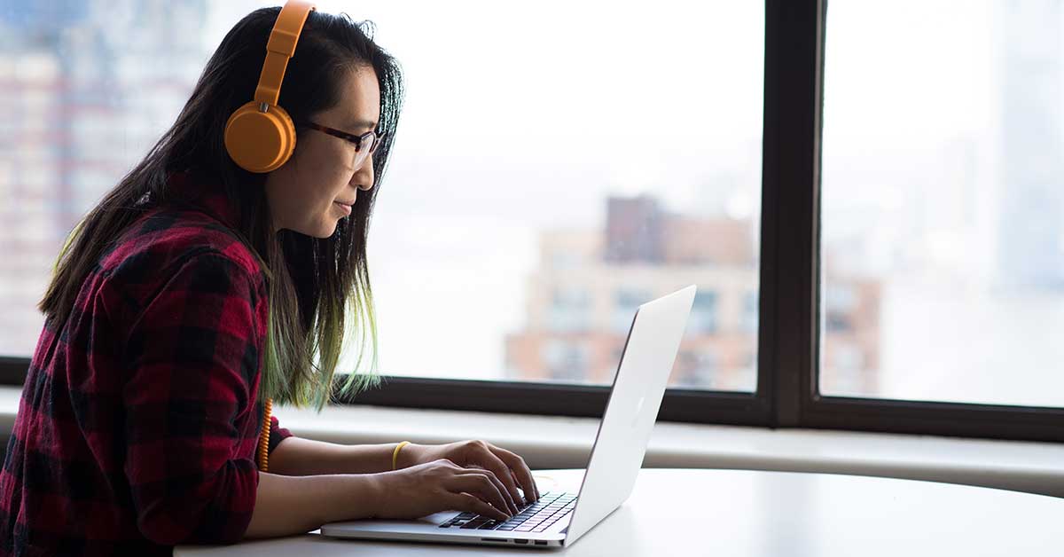 photo of woman sitting at a desk wearing headphones typing on a laptop