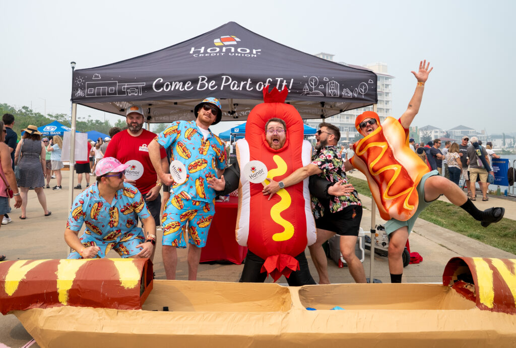 People in colorful hot-dog costumes posing under a black Honor Credit Union tent at an outdoor festival with a hot-dog-themed obstacle course in the foreground. | Honor Credit Union | Honor Credit Union