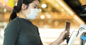 image of a woman wearing a medical face mask holds her phone up to a credit card terminal to make a touchless payment | Honor Credit Union