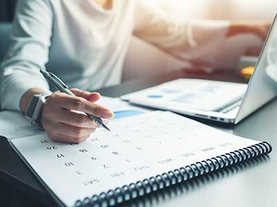 image of a person sitting at a desk writing on a calendar while looking at a laptop screen