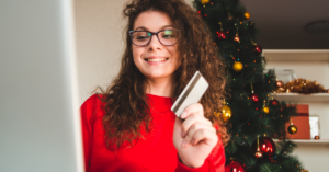Young woman with glasses shopping online at a computer, holding a credit card, Christmas tree in the background. | Honor Credit Union