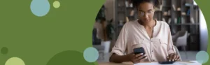 woman looking at her phone while sitting at a table with a calculator and paperwork | Honor Credit Union