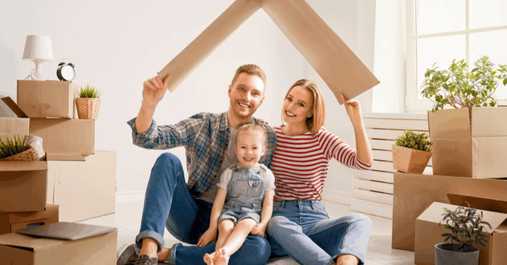 a young family sitting on the floor with a cardboard box above their heads pretending it is a roof smiling and happy to move into their new home