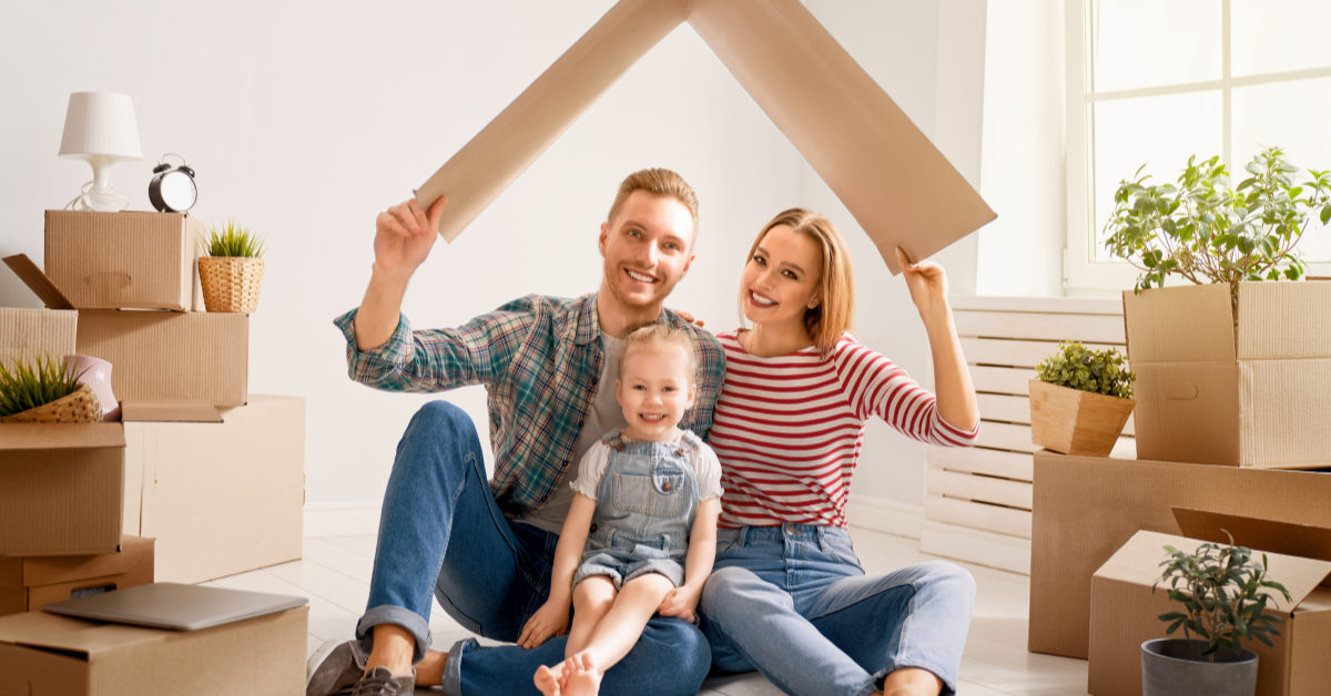 a young family sitting on the floor with a cardboard box above their heads pretending it is a roof smiling and happy to move into their new home