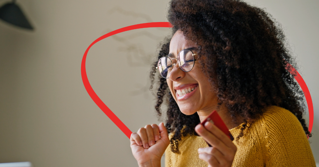 Smiling woman with curly hair and glasses holds a red card, celebrating a purchase or reward. | Honor Credit Union