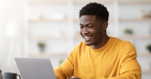 Young man in a mustard sweater smiling while using a laptop in a bright home office. | Honor Credit Union