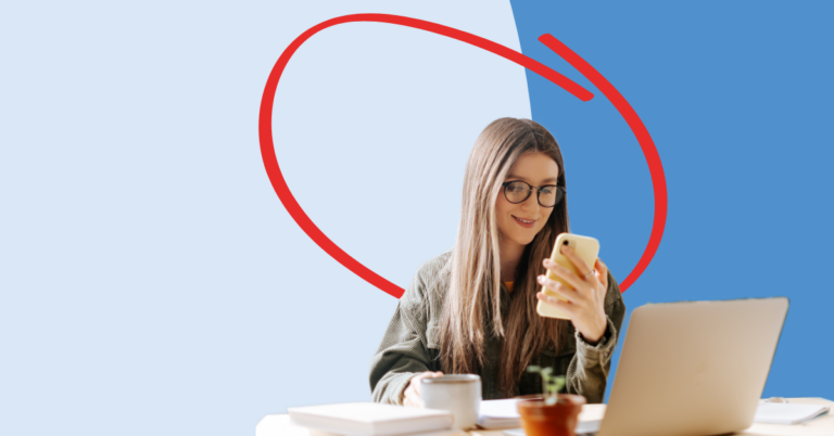 Young women looking at a cell phone, sitting at a desk with a laptop and coffee.