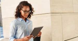 Woman in a light blue button-down shirt with glasses uses a tablet outdoors beside beige stone walls. | Honor Credit Union