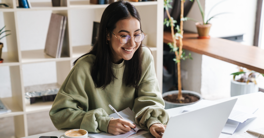 Young woman with glasses studying at a desk, writing notes beside a laptop and coffee cup in a bright room. | Honor Credit Union