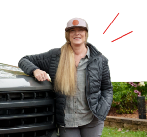 Woman in a gray shirt and black puffer jacket leaning on the front of a dark SUV, smiling outdoors, wearing a cap. | Honor Credit Union