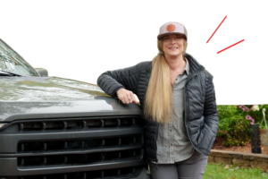 Woman with long blonde hair, cap, and jacket leaning on the front of a green SUV, smiling. | Honor Credit Union
