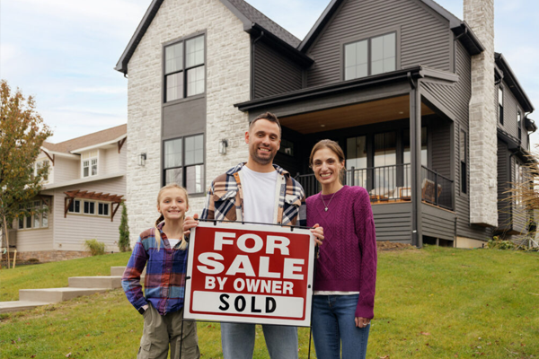 Family and child pose with a 'For Sale by Owner—Sold' sign in front of a two-story house.
