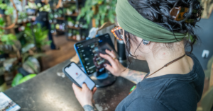 Person with a green headband and glasses uses a tablet and smartphone at a plant-filled cafe counter, concentrating on the screens. | Honor Credit Union