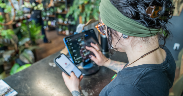 Person with a green headband and glasses uses a tablet and smartphone at a plant-filled cafe counter, concentrating on the screens. | Honor Credit Union | Honor Credit Union