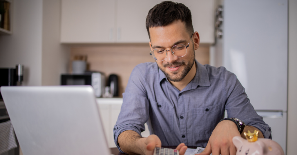 Man with glasses working on a laptop in a kitchen, using a calculator with a piggy bank nearby on the desk | Honor Credit Union