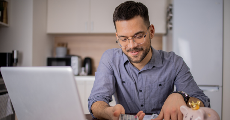 Man with glasses working on a laptop in a kitchen, using a calculator with a piggy bank nearby on the desk | Honor Credit Union | Honor Credit Union