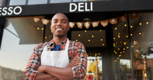 Smiling man in a plaid shirt and white apron stands with arms crossed outside a deli storefront at dusk. | Honor Credit Union