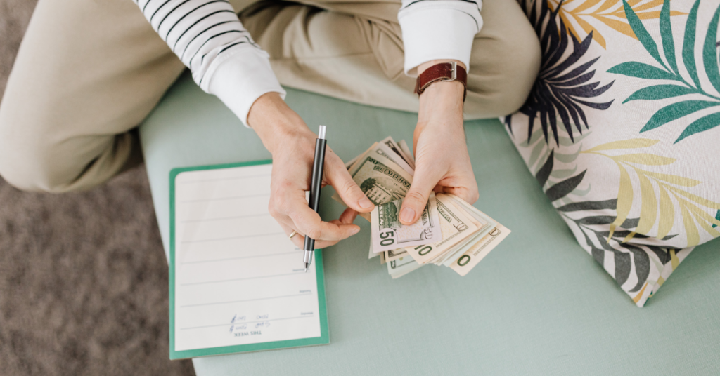 Person sitting on a mint-green cushion counts cash ($50 bills) while writing on a clipboard with a pen, suggesting budgeting or expenses planning. | Honor Credit Union