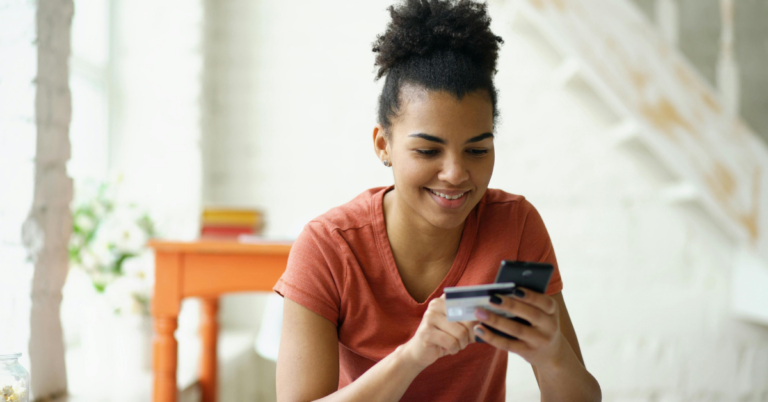 Smiling woman in an orange shirt sits indoors, holding a phone and a credit card for online shopping or mobile payment. | Honor Credit Union | Honor Credit Union