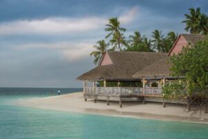 Thatched-roof beach pavilion on stilts along a white-sand shoreline with turquoise water and palm trees in a tropical coastal scene. | Honor Credit Union
